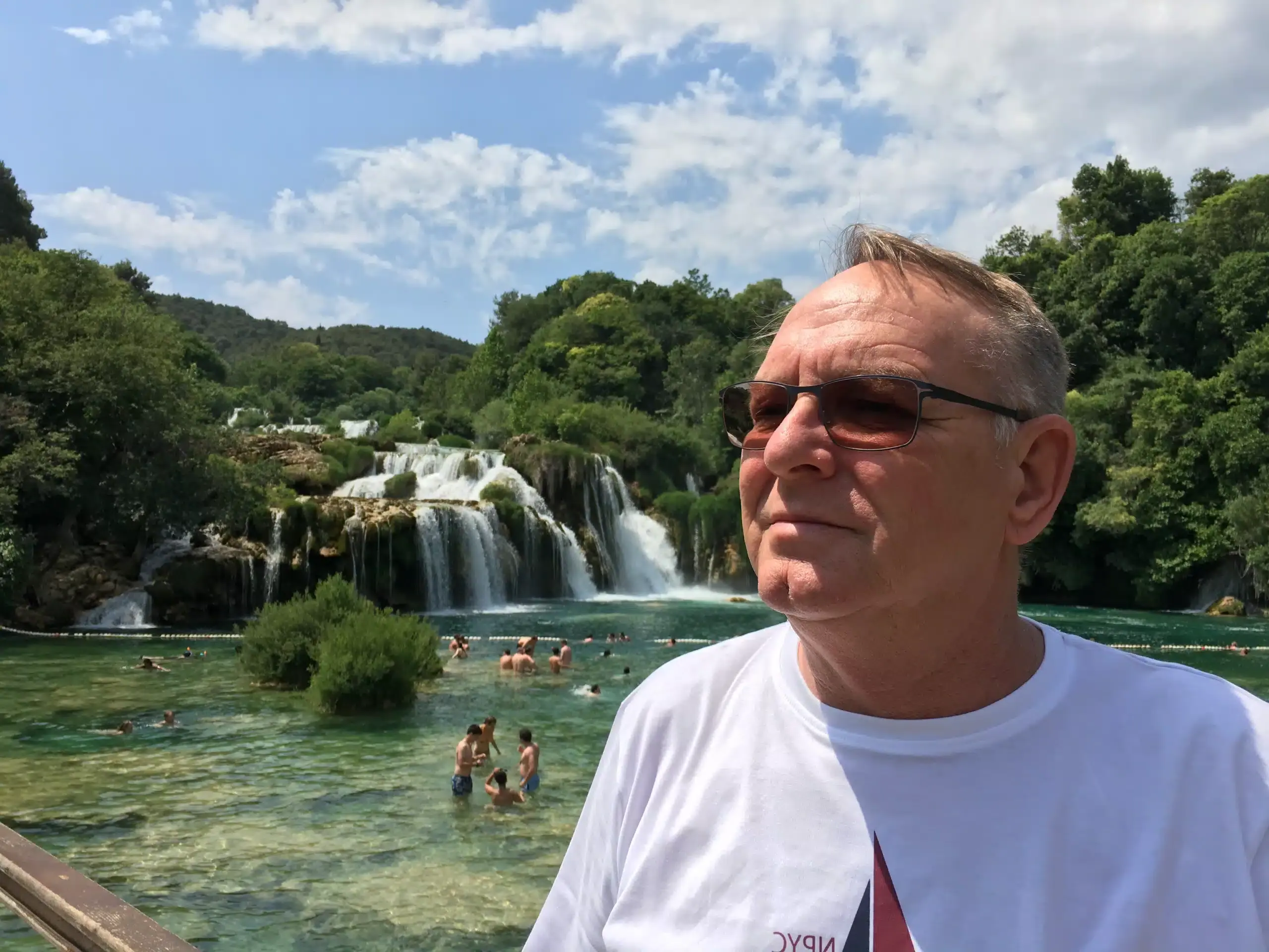 Ein lächelnder Mann mit Sonnenbrille steht im Vordergrund, während im Hintergrund Menschen in einem klaren Fluss mit Wasserfällen baden und die üppige, grüne Natur genießen.