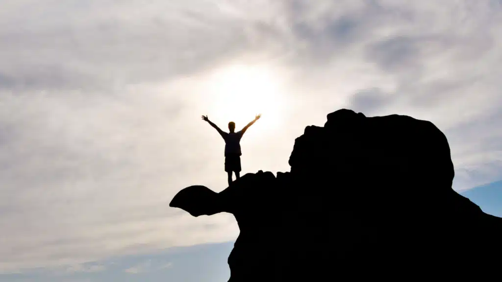 Silhouette eines Mannes auf einem Felsen mit erhobenen Armen im Sonnenlicht als Symbol für Neustart im Leben mit 50