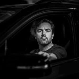html Kopieren Schwarz-Weiß-Porträt eines konzentrierten Mannes am Steuer eines Autos, der mit ernstem Blick nach vorne schaut – ausdrucksstark und dynamisch.