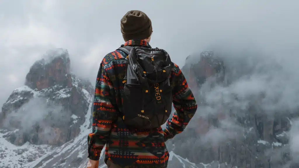 Ein Wanderer mit Rucksack steht vor einem beeindruckenden Bergpanorama mit Wolken – Symbol für Abenteuer, Freiheit und die Schönheit der Natur.